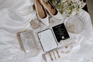 Wedding details flatlay with white shoes, invitation cards, delicate jewelry and baby's breath flowers on white fabric.