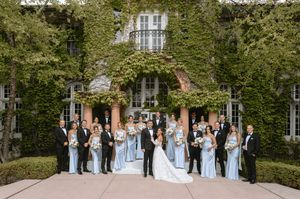 Large wedding party poses before ivy-covered mansion, bridesmaids in light blue gowns with groomsmen in black tuxedos.