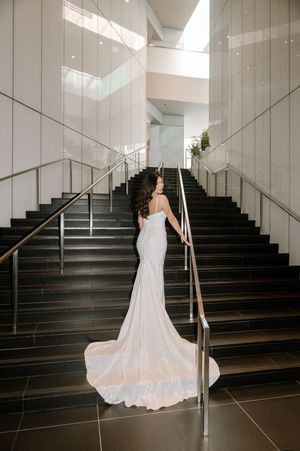 Bride in elegant white mermaid gown with long train standing on modern staircase, viewed from behind.