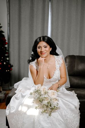 A bride in a white lace wedding dress poses with a delicate bouquet of white flowers against a light background.