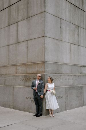A couple poses at an urban concrete building corner, the bride wearing a tea-length dress and holding a bouquet.