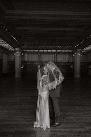 Black and white photo of bride and groom sharing a kiss under a veil in an elegant ballroom.