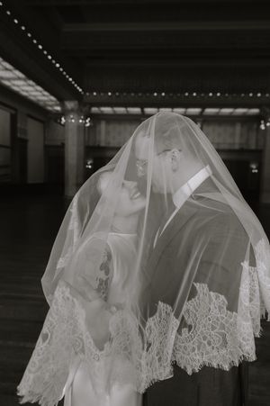 Black and white wedding photo of bride and groom sharing a kiss under a lace veil in an elegant venue.
