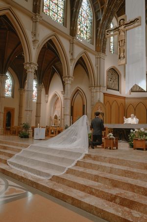 Couple kneeling at church altar during Catholic wedding ceremony with long bridal veil trailing down marble steps.