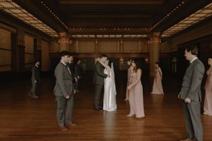 Wedding party stands in circle during first dance in elegant ballroom with warm string lights overhead.
