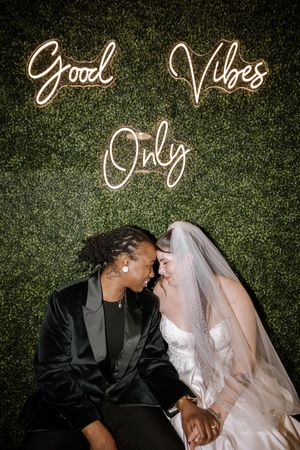 Newlyweds share an intimate moment under glowing 'Good Vibes Only' neon sign against a green wall backdrop.