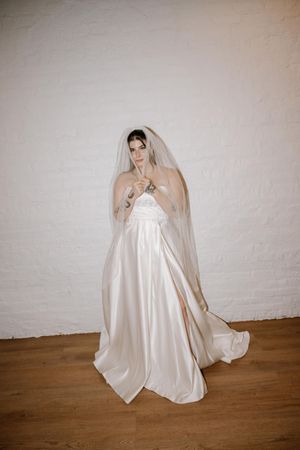 A bride in an elegant white satin wedding gown and long veil poses gracefully against a light colored wall.