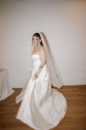 A bride in a flowing white satin wedding gown and long veil poses against a light colored wall.