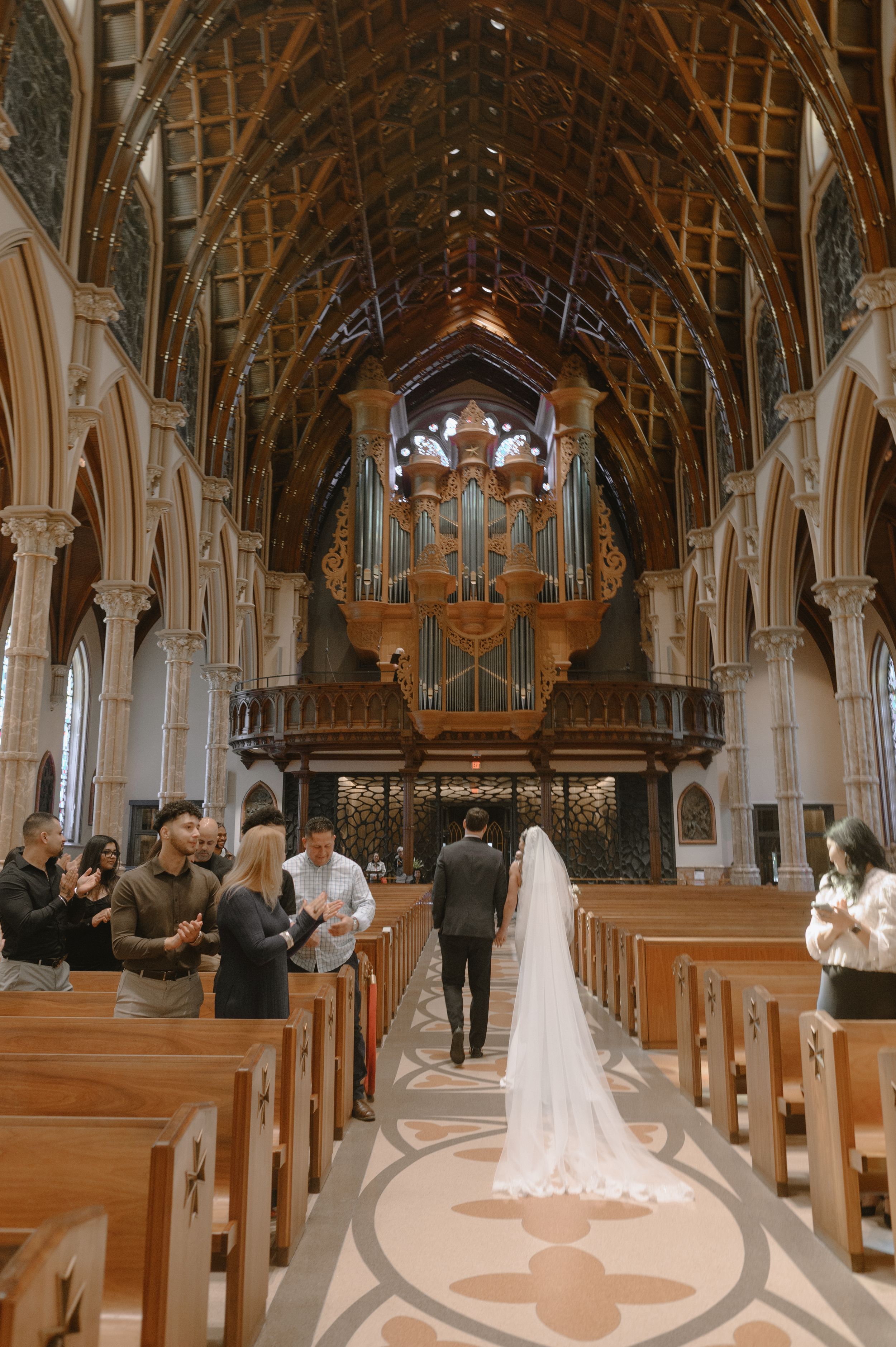 Bride walking down the aisle of a grand Gothic church with vaulted ceilings and a large pipe organ.