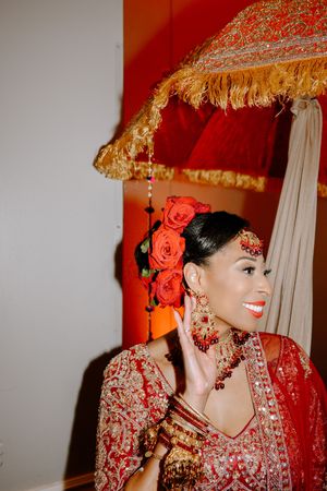 Bride in red lehenga with rose hair accessories smiles under golden ceremonial umbrella at Indian wedding.