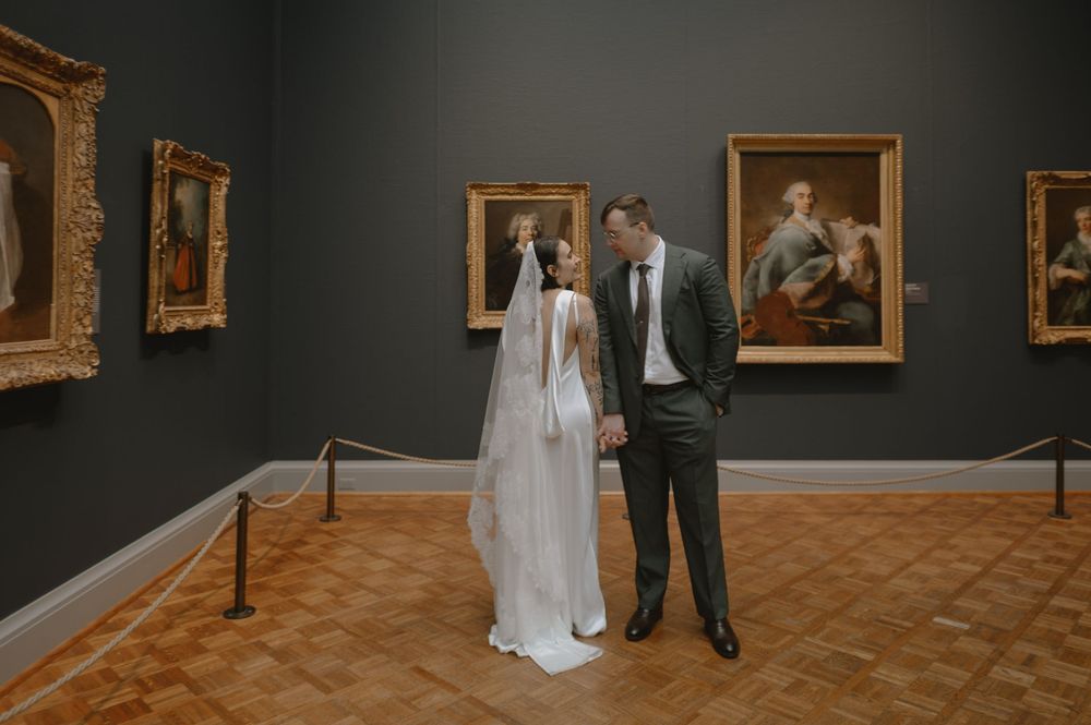 Bride and groom share romantic moment in art museum gallery surrounded by classic portrait paintings.