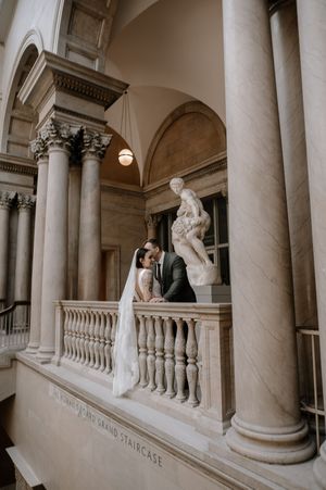 Bride and groom share a romantic moment on the Grand Staircase balcony of a classical marble building with columns.