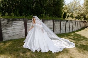 Bride in flowing white wedding gown and long veil poses gracefully in garden setting with rustic wood fence backdrop.