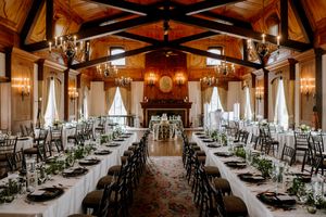 Elegant wedding reception hall with long tables, white linens, greenery centerpieces, chandeliers, and a grand fireplace.