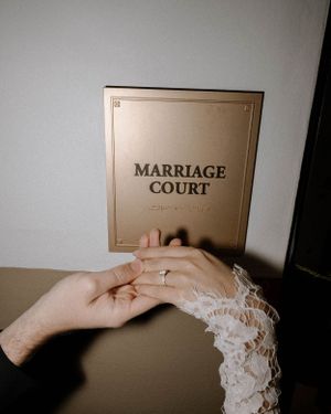 Hand wearing lace sleeve and wedding ring holds a Marriage Court document in moody lighting.