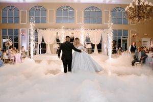 A couple dances on a misty white floor beneath an elegant white wedding canopy in a grand ballroom with arched windows.