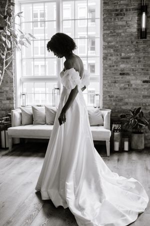 A graceful silhouette in a flowing white wedding gown stands by a window in an industrial-chic loft space with exposed brick.