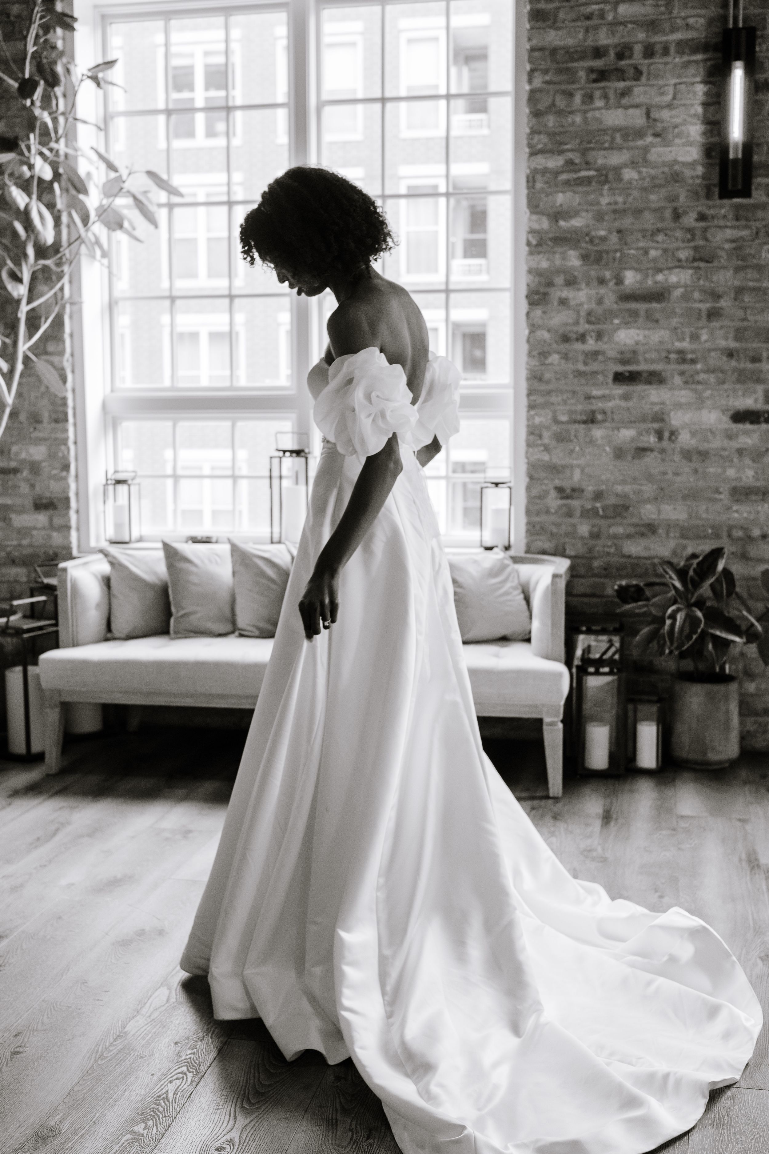 A graceful silhouette in a flowing white wedding gown stands by a window in an industrial-chic loft space with exposed brick.