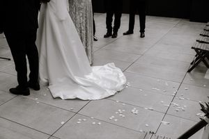 Wedding guests throwing white flower petals on the ground around a bride's flowing dress at an indoor ceremony.