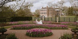 Bride in white gown poses in formal garden with pink tulips, ornate hedges, and Georgian manor backdrop.