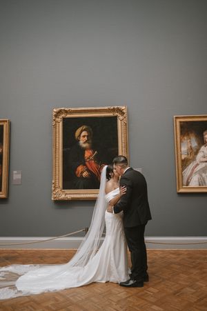 Bride and groom share a kiss in an art museum gallery, with classic gold-framed portraits on gray walls.