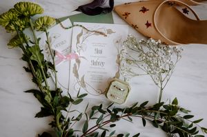 Wedding flat lay with floral invitation suite, ring box, baby's breath, greenery, and tan heels on marble surface.