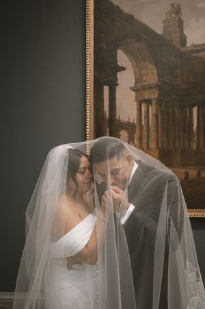 Bride and groom sharing an intimate moment under a veil in front of a classical painting.
