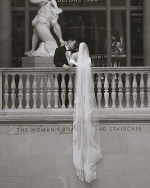 Black and white wedding photo of bride and groom kissing on ornate balcony with flowing veil and classical sculpture.