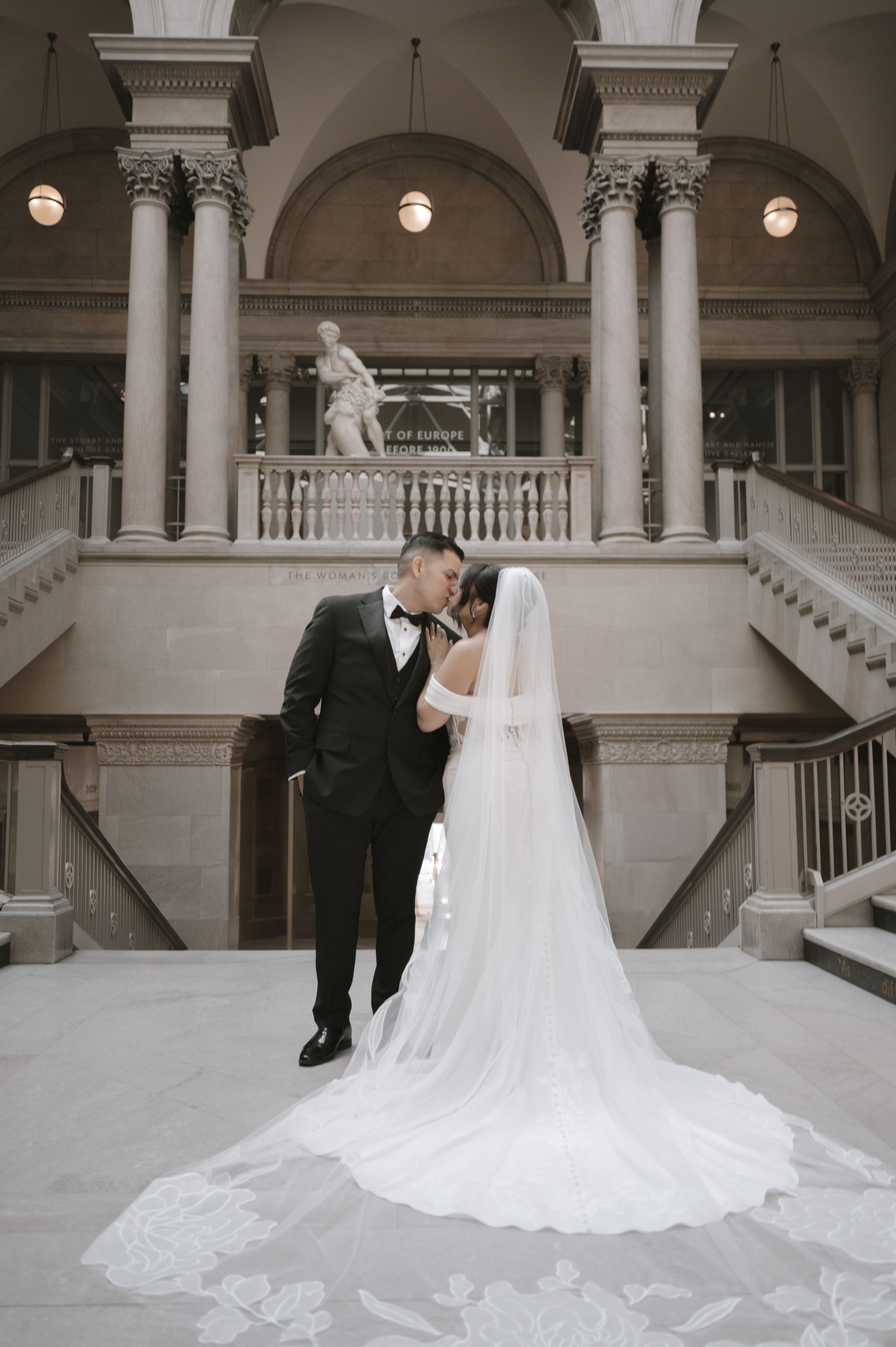 Bride and groom share romantic moment in elegant grand hall with marble columns, sweeping staircase, and cathedral veil.