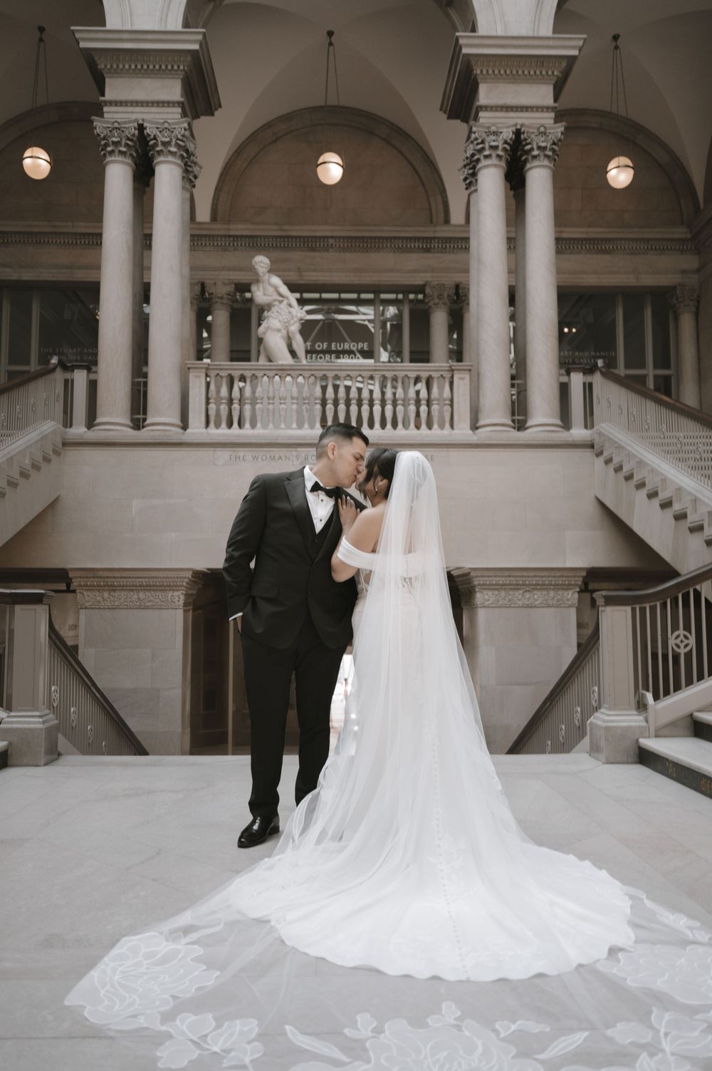 Bride and groom share romantic moment in elegant grand hall with marble columns, sweeping staircase, and cathedral veil.