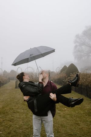 Two people dance playfully with umbrellas in a misty field on an overcast day.