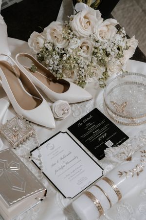 Elegant wedding details featuring white roses, pearl accessories, bridal shoes, and black and white invitation cards.