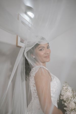 A bride in a white lace wedding dress with a flowing veil poses elegantly in a light-filled setting.
