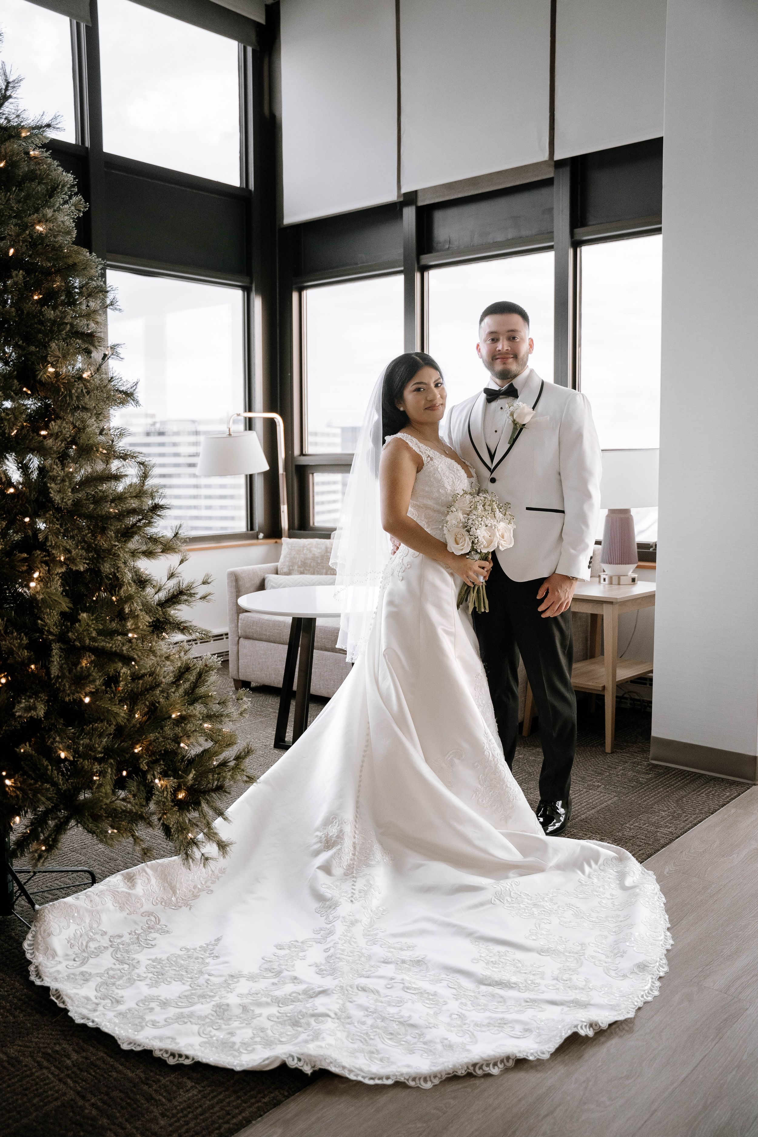 A couple in elegant wedding attire poses in a modern room with large windows and a decorated Christmas tree.