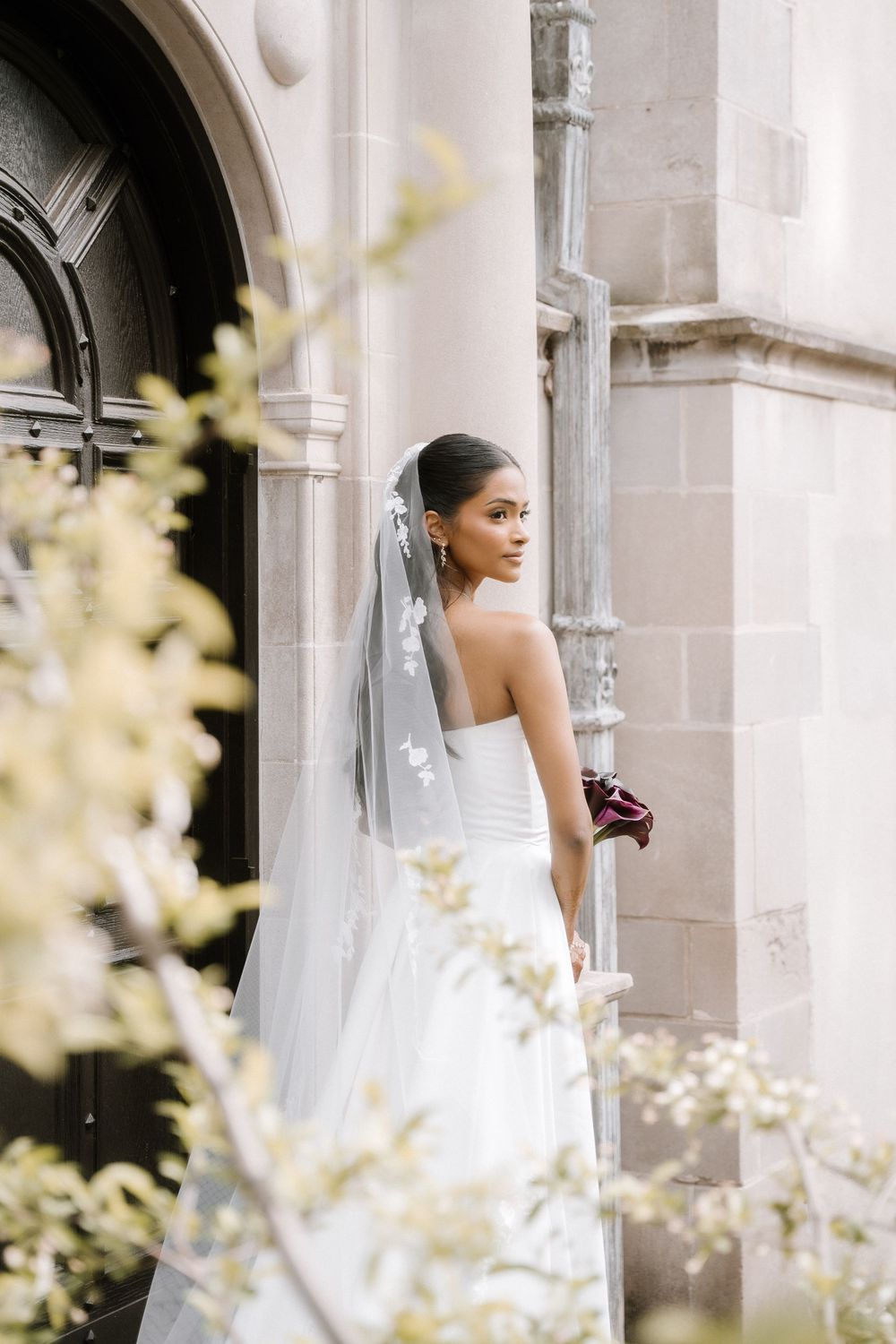Elegant bride in strapless white gown and veil stands by stone church entrance, holding bouquet.
