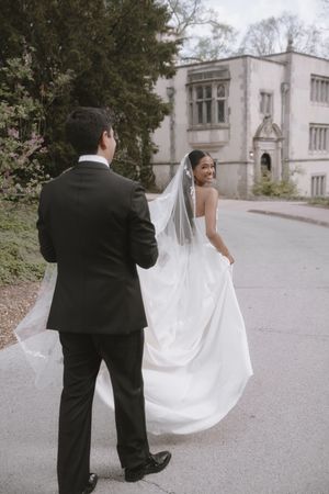Bride in white gown and veil smiles back at groom in black suit outside elegant stone mansion.