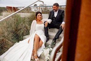 A couple in wedding attire sits together on outdoor steps surrounded by coastal beach grass.