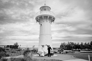 A black and white photograph of a couple embracing near a tall white lighthouse under dramatic cloudy skies.