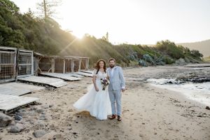 A couple in wedding attire walking along a beach with wooden cabanas.