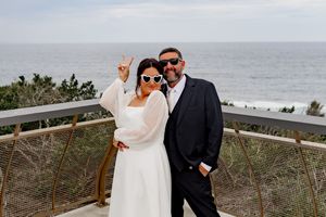 Couple in formal wedding attire poses together on a scenic coastal boardwalk overlooking the ocean.