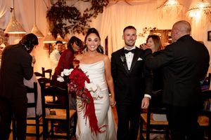 Bride in white gown holding red bouquet walks with groom in black tuxedo at elegant indoor wedding reception.