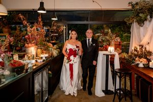Bride in white gown holding red bouquet walks with suited man through elegantly decorated wedding venue.