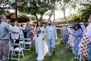 Outdoor wedding ceremony with guests seated on white chairs among trees.