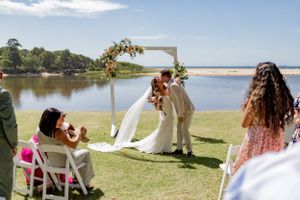 Wedding ceremony setup by a lake with white chairs and floral arch against blue sky.