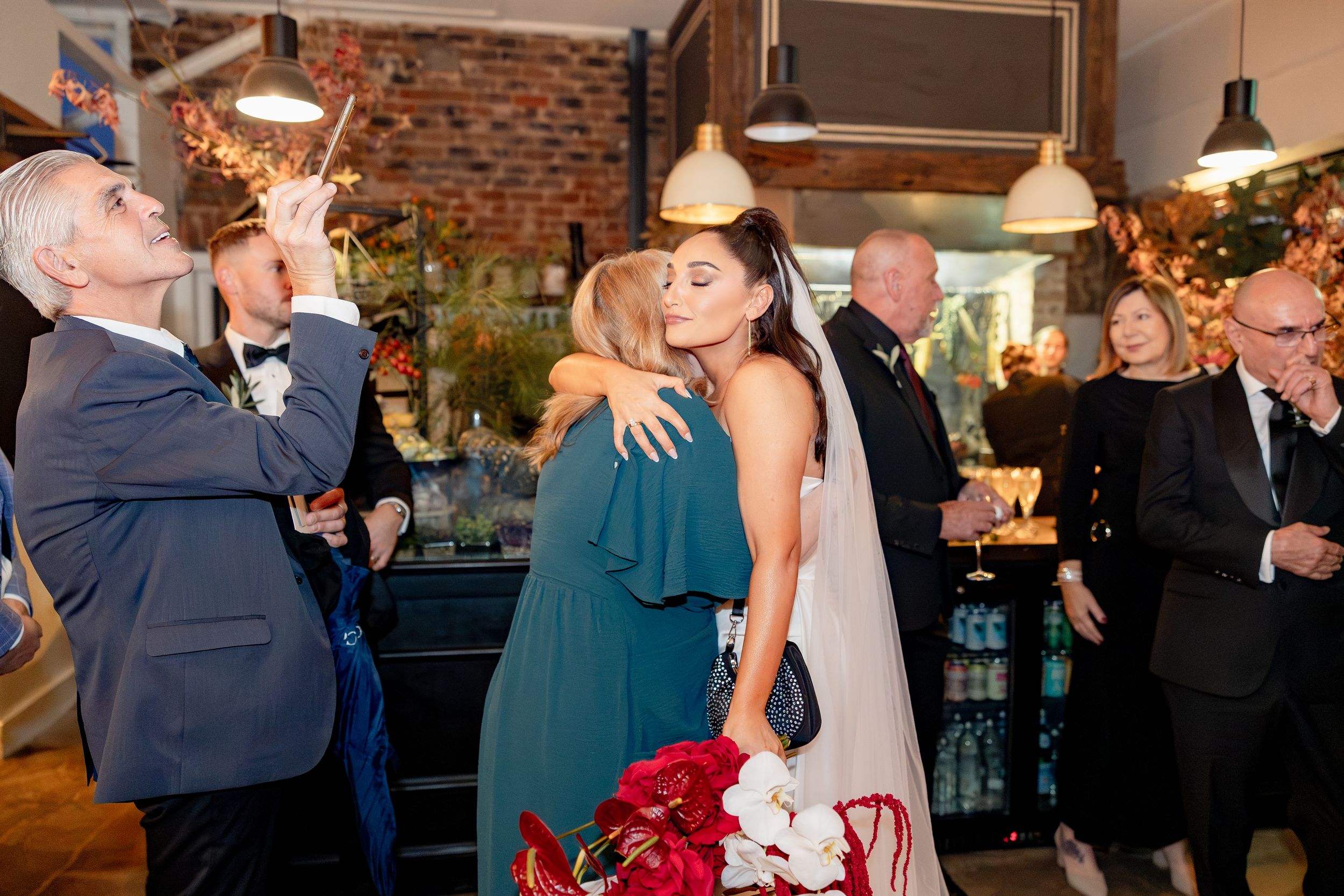 Bride in white gown hugging bridesmaid in teal dress at wedding reception while guests celebrate around them.
