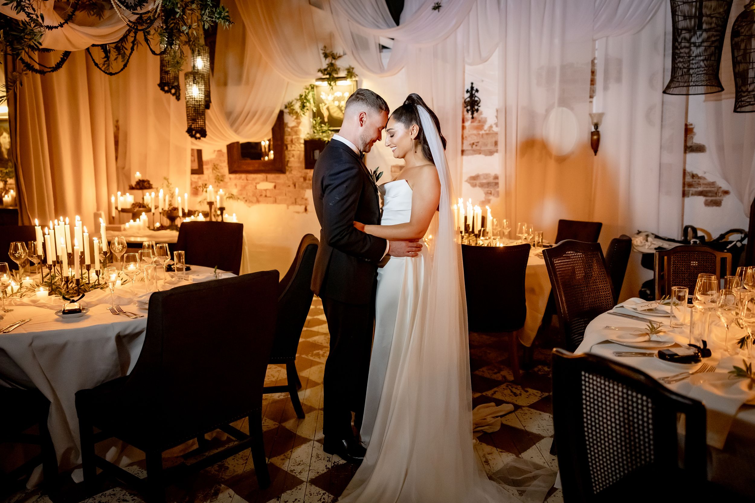 Bride and groom share a romantic kiss in a warmly lit wedding reception venue with candlelit tables.