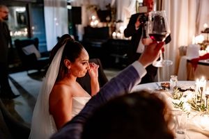 Bride in white gown and veil smiles as guest raises red wine glass during elegant candlelit wedding reception.