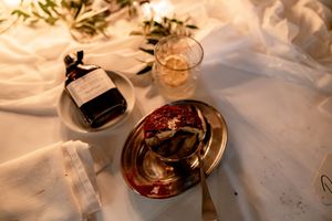 Elegant table setting with pomegranate, glass bottle, amber drink, and olive branch on white linen.