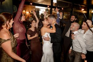 Bride and groom share a kiss surrounded by cheering guests holding sparklers at a lively wedding reception.