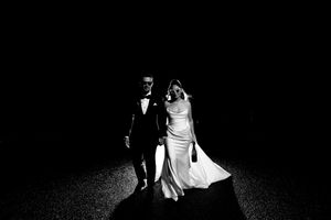 Black and white wedding photo of bride and groom walking together at night, illuminated against dark background.